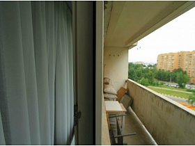 стул, стол, подушки и пакеты в углу открытого балкона современной однокомнатной квартиры из открытой двери гостиной с белой гардиной на окне