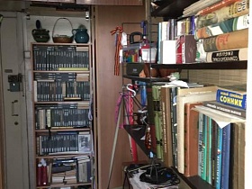 обувь на коврике перед этажеркой с вазочками, корзиной на верху и книгами на полках у входной двери в прихожую трехкомнатной квартиры