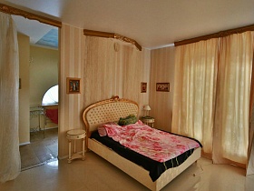 гримерные круглые столики по обе стороны бежевой кровати с розовым постельным , иконы на стене, бежевые шторы на окне бежевой спальни художественной дачи в сосновом лесу