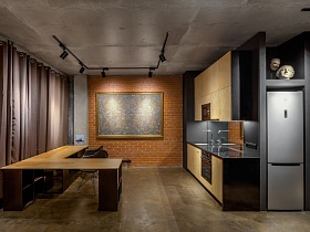 встроенный серебристый холодильник, бежевая кухня ,бежевый угловой стол у окон с коричневыми шторами в стильной студии с черными софитами на бетонном потолке