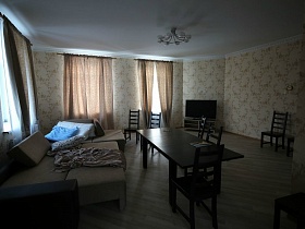 разложеннй бежевый с коричневым диван с подушками, стол со стульями, плоский телевизор, люстра на белом потолке светлой гостиной кирпичного двухэтажного дома под съем