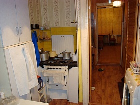 полотенца, кухонный инвентарь, металлическая угловая полка на стене с желтой плиткой в нише с газовой плитой и кастрюлями на встроенных шкафах небольшой кухни с белым шкафом и стиральной машинкой двухкомнатной квартиры бабушки советского времени