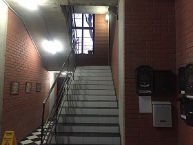 небольшие картины на стене из красного кирпича на первом этаже с шахматной плиткой на полу и  лестницей с перилами между этажами