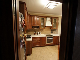 коричневая мебельная стенка с белой столешницей, холодильник на полу с бежевой плиткой кухни современной трехкомнатной квартиры государственного служащего