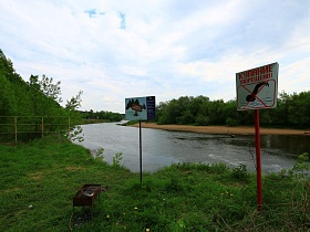 предупредительные плакаты на металлических стойках, мангал и оградительная сетка на берегу реки с песчанным пляжем летом