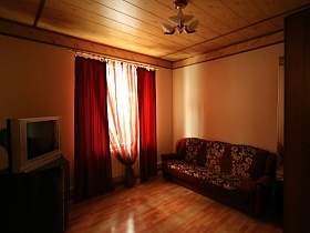 красные шторы и красная гардина на окне, телевизор на тумбочке, раскладной диван в гостевой спальне пустого двухэтажного съемного дома в сосновом лесу