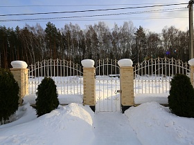 зеленый кипарис на заснеженом участке с расчищенной дорожкой к воротам в резном белом металическом заборе с колонами вокруг современного загородного коттеджа