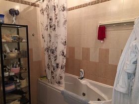 светлая цветная шторка над белой ванной, косметические препараты на полках стеклянного шкафчика в ванной комнате с двухцветной бежевой плиткой на стенах