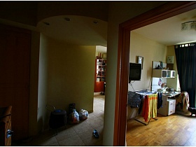 телевизор на ножке на стене, сушилка для белья, письменный стол со шкафчиками, темно-серая  кошка  на полу с линолеумом в спальне через открытую дверь из прихожей двухкомнатной квартиры с видом на Москва-сити
