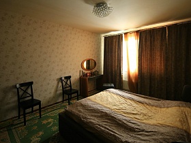 коричневые тяжелые шторы на большом окне спальной комнаты с люстрой на белом потолке, зелеными коврами на полу простой современной трехкомнатной квартиры