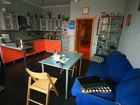 квадратные часы над входной дверью кухни с ораньжевой мебельной стенкой, синим мягким диваном, обеденным столом и большим шкафом с посудой трехкомнатной квартиры