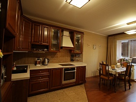 коричневая квадратная плитка на рабочей поверхности коричневой кухни, обеденный стол и стулья со спинками у застекленной лоджии, совмещенной с кухней трехкомнатной квартиры государственного служащего