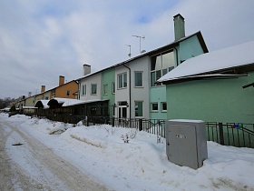 сугробы снега у металлического забора вокруг одноуровневых жилых домов, разных цветов, с навесами над крыльцом