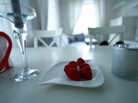 красно белая салфетница в виде сердечка, свеча в стеклянном подсвечнике, сочная клубника на белой тарелочке и стакан на поверхности белого обеденного стола на кухне