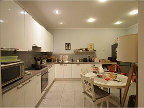 белый круглый стол, белая мебельная стенка с серой столешницей на полу с белой плиткой светлой кухни просторной современной квартиры
