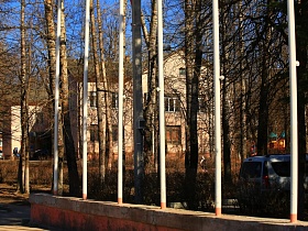 двухэтажный жилой дом с треугольной крышей в просвете густо высаженных стволов лиственных деревьев за флагштоком в городке Сычево