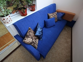 голубой мягкий диван с разноцветными квадратными подушками на сером полу застекленного балкона современной дизайнерской трехкомнатной квартиры