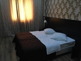большая кровать с белыми подушками, постельным и коричневым покрывалом у высокого изголовья, прикроватные коричневые деревянные тумбочки в номере с цветочными и полосатыми обоями