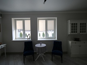 белый прямоугольный столик у стены с кондиционером, белый навесной шкаф со стеклянными дверцами над тумбой, вазочка с цветами на круглом столе со скатертью, синие кресла у окна с ролетами на кухне загородного дома