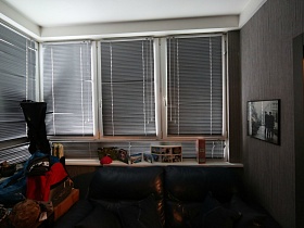 серый мягкий диван с подушками у окон просторной лоджии с закрытыми жалюзи на окнах, серыми стенами и белым потолком молодежной квартиры в современном мегаполисе
