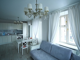 белые шторы на больших окнах зонированной светлой комнаты с белой мебелью и белыми плафонами подвесной люстры на белом потолке двухкомнатной квартиры