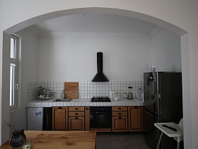 коричневая кухня с черной вытяжкой на белой стене над газовой плитой,серый холодильник, детский стульчик для кормления напротив узкого окна в кухне зонированной комнаты современной скандинавской квартиры