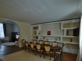 большой арочный дверной проем в столовую с длинным коричневым деревянным столом со стульями на большом двухцветном ковре и белыми полками у стены современного кирпичного дома