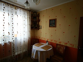 чайник, салфетки на круглом столе с белой скатертью, картина на стене,иконы на открытых полках в углу кухни с белой гардиной на окне с балконой дверью яркой трехкомнатной квартиры с комнатой бабушки