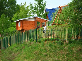 деревянный дом с синей крышей, различными постройками за цветным металлическим забором на крутом берегу реки с островками и изгибом