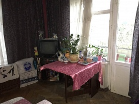 черный телевизор на открытой тумбочке в углу сиреневой гостиной, комнатные цветы, кувшин, сахарница и поднос на раскладном столе с сиреневой скатертью у окна с балконной дверью