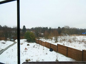 вид из окна дома на большой участок под снегом за деревянным забором и огромный пустырь