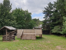 деревянный бревенчатый колодец с треугольной крышей, открытая загороженная трибуна со ступенями на большом дворе с высокими зелеными соснами за деревянным забором в небольшой деревне