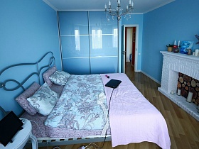 цветы и свечи на белом декоративном камине,шкаф для одежды с матовыми голубыми стеклянными дверцами ,большая кровать в спальне с голубыми стенами уютной женской квартиры в новостройке