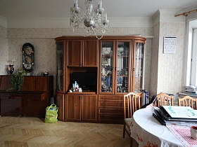 большие настенные часы над коричневым пианино , коричневая стенка с посудой под стеклом в гостиной квартиры сталинки