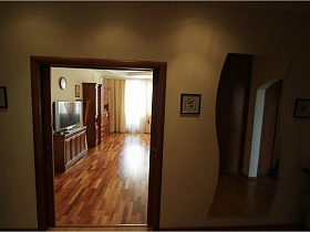 волнистое зеркало на стене с маленькими картинами в прихожей у открытой двери в гостиную с телевизором на комоде и мебельной стенкой простой средней двушки с балконами