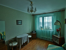 трельяж, плетенные кресла у овального стола с белой скатертью, люстра, стилизованная под свечи в бирюзовой комнате яркой трехкомнатной квартиры с комнатой бабушки