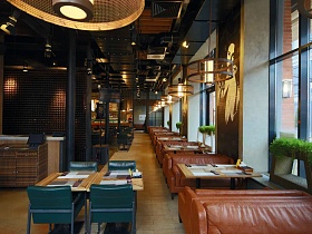 ряд прямоуголных столиков с коричневыми мягкими диванчиками у больших окон и квадратные столы с синими мягкими стульями по центру уютного крафтового ресторана