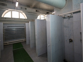 открытые душевые кабины с белым кафелем на стенках и перегородках в просторной комнате с арочными полукруглыми окнами под потолком и системой отопления на стене