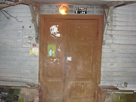 уличный фонарь над окрашенными входными деревянными дверьми кирпичного дома с отбитой плиткой на фундаменте
