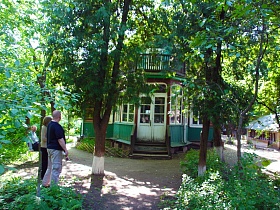 ступени крыльца с перилами к входным белым дверям деревянной художественной дачи-музей среди высоких зеленых деревьев на участке за зеленым забором