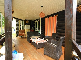 темная плетенная мебель со столиком на открытой веранде со светлым потолком темной деревянной дачи-сруб директора