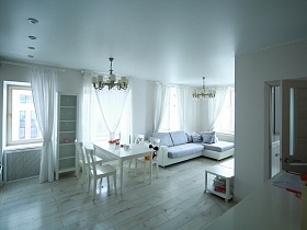 обеденный стол со стульями и угловой мягкий диван у больших окон с белой гардиной зонированной комнаты стильной современной квартиры в новостройках