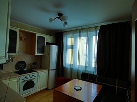 белая стенка с затемненными стеклами, угловой темный диван с белыми полосками у раскладного стола и темные шторы на окне кухни большой трехкомнатной квартиры после переезда