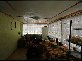 клеенка в клетку на длинном столе с кастрюлями, диван с подушками и белый холодильник в углу веранды с окнами на всю стену на даче
