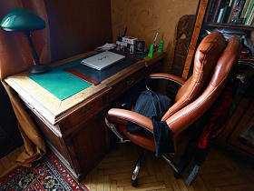 ноутбук, фотографии, письменные принадлежности, настольная лампа с зеленым абажуром на старинном деревянном столе с зеленой поверхностью в спальне трехкомнатной советской квартиры