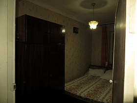 коричневый полированный шкаф для одежды с антресолью, люстра с цветами над двумя односпальными кроватями с белыми подушками в спальной комнате молодой советской семьи