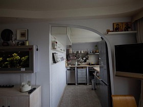 плоский телевизор, иконы на белой настенной полке с одной стороны,настенный аквариум с другой арочного дверного проема из гостиной на кухню стильной трешки