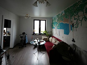 круглая стеклянная поверхность журнального столика на трех ножках у бардового мягкого дивана с подушками в гостиной простой разрисованной трехкомнатной квартиры