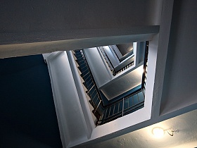 вид снизу на белые лестничные пролеты редкой винтовой лестницы с металлическими перилами и деревянными поручнями в подъезде жилого девяти этажного дома с синими стенами