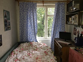 зеленая листва деревьев из окна с голубыми шторами спальной комнаты с нежно-розовым постельным с розами на деревянной кровати спальной комнаты двухкомнатной квартиры